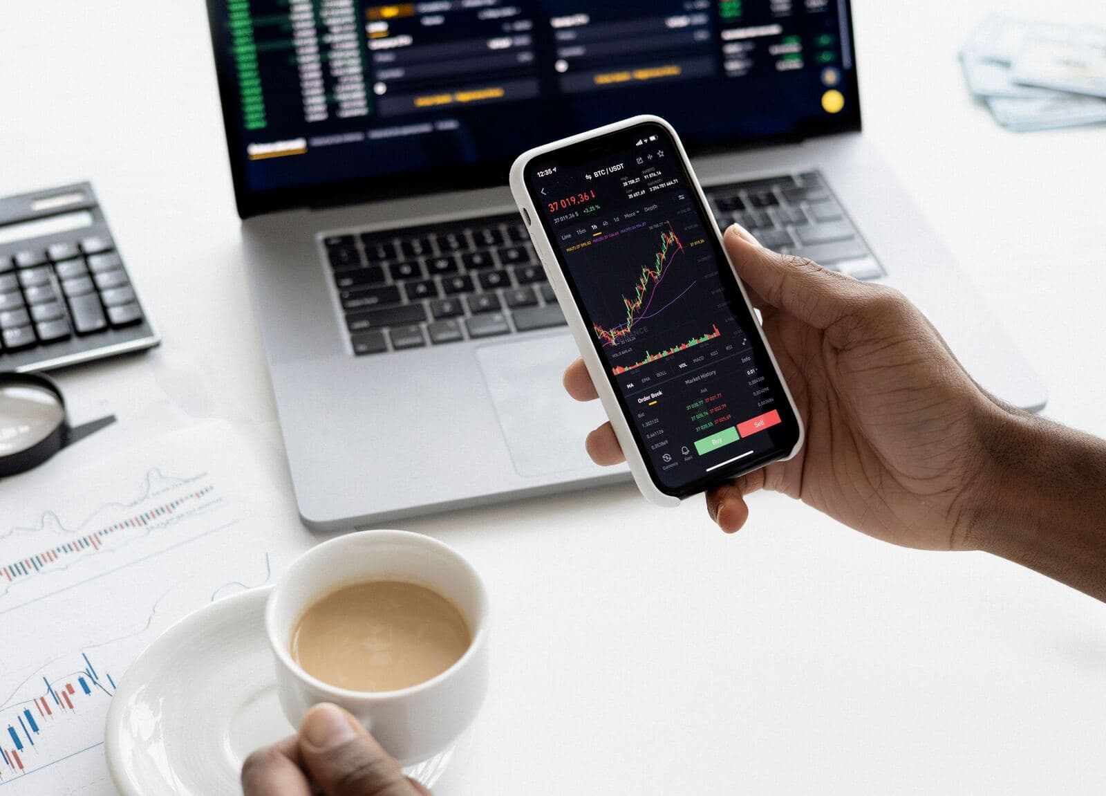 A person checking a stock chart on a phone beside a laptop and papers